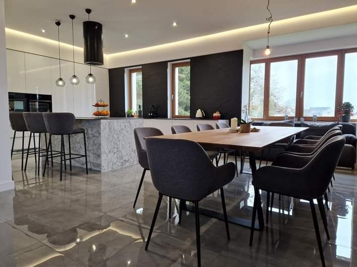 Jasne wnętrze jadalni z otwartą kuchnią; wyspa kuchenna z marmurowym wykończeniem, grafitowe krzesła tapicerowane wokół drewnianego stołu na metalowej podstawie, błyszcząca podłoga z płytek, duże...