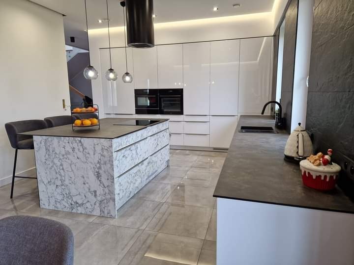 Nowoczesna kuchnia z wyspą wykończoną marmurem, wiszące lampy kuliste, białe szafki bez uchwytów i szary blat.