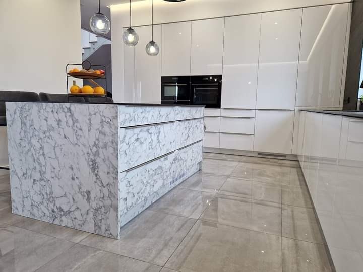 Nowoczesna kuchnia z białymi, lakierowanymi szafkami, wyspą z marmurowym wzorem i szarymi płytkami podłogowymi, oświetlona trzema wiszącymi lampami kulami.