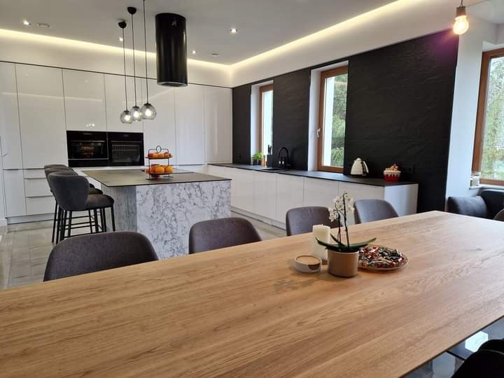 Jasna, nowoczesna kuchnia z białymi szafkami, wyspą z marmurowym wykończeniem i czarnym panelem ściennym. Na pierwszym planie drewniany stół z dekoracjami.