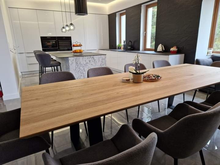 Jasny, nowoczesny salon z widoczną jadalnią: duży drewniany stół na czarnych nogach, szare krzesła, w tle biała kuchnia z wyspą i czarnymi elementami dekoracyjnymi.