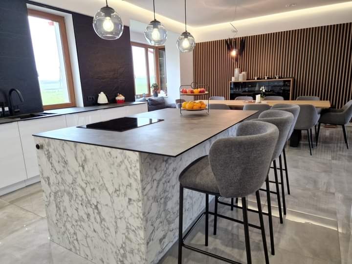 Nowoczesna kuchnia z wyspą wykończoną marmurem, szare hokery barowe, drewniana ściana dekoracyjna w tle, oświetlenie punktowe i duże okna wpadające do wnętrza.