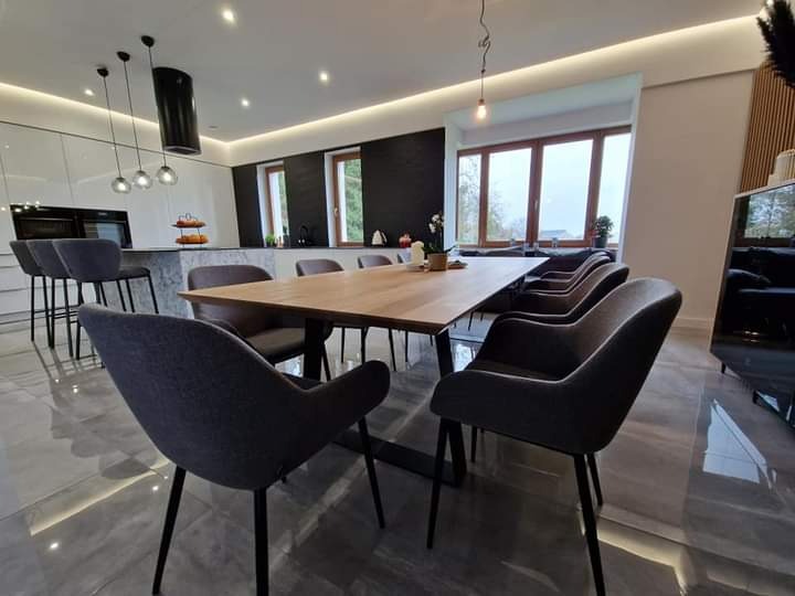 Nowoczesna jadalnia z długim drewnianym stołem na metalowych nogach, otoczonym szarymi krzesłami z podłokietnikami, w tle otwarta kuchnia z wyspą i hokerami, całość w jasnej kolorystyce z czarnymi...