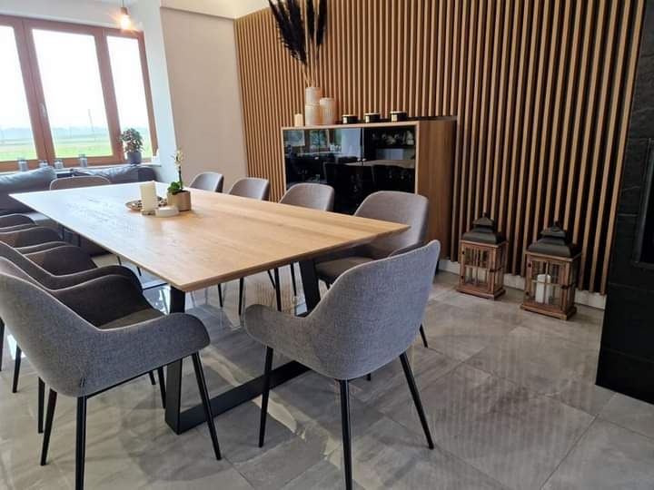 Jasna, dębowa jadalnia z szarymi krzesłami na czarnych nogach, dekoracyjna drewniana ściana z pionowych listew, szare płytki podłogowe i widok na zewnątrz przez duże okno.