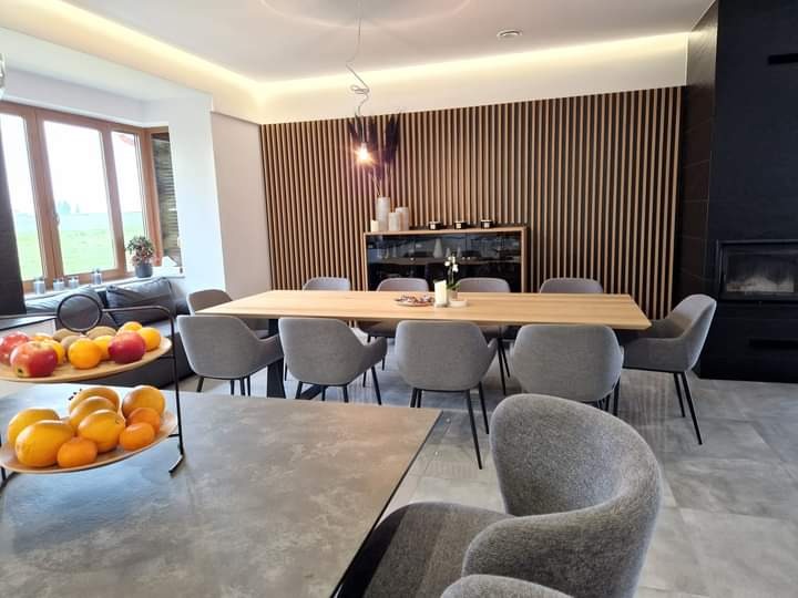 Jasna jadalnia w nowoczesnym domu z drewnianą ścianą z pionowych lameli, długim drewnianym stołem otoczonym szarymi krzesłami, owocami w dwupoziomowej paterze i widokiem na zieleń za oknem.