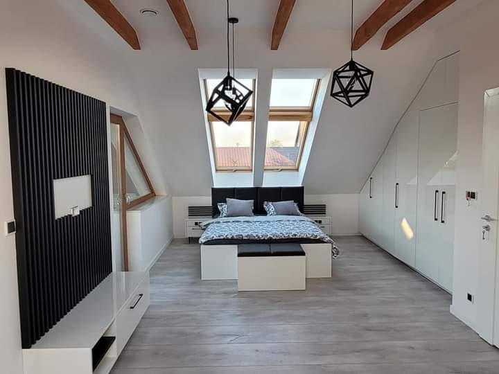 Pokój na poddaszu ze skośnym sufitem i drewnianymi belkami, łóżko z szarą pościelą, szafa w zabudowie, czarna dekoracyjna ściana z białym prostokątem, dwie lampy wiszące o geometrycznym kształcie.