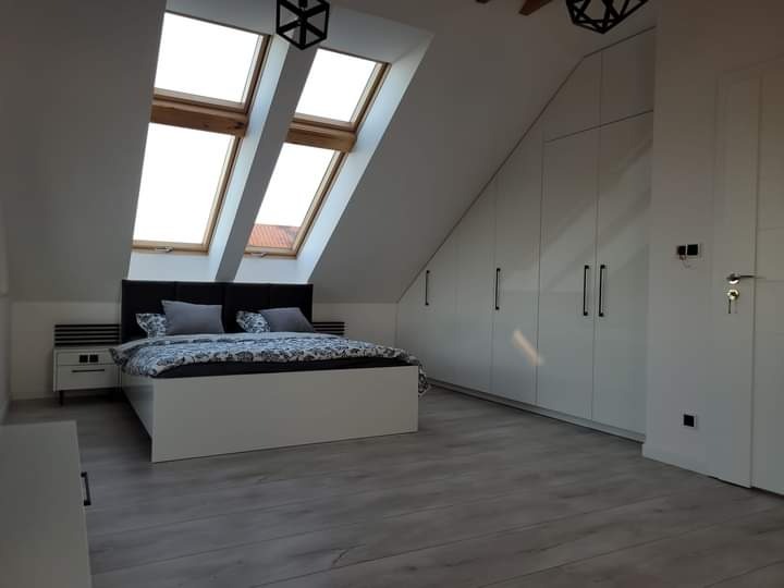 Pokój na poddaszu z łóżkiem, szafą w zabudowie i oknami dachowymi, utrzymany w tonacji bieli i szarości, z drewnianą podłogą i czarnymi lampami.