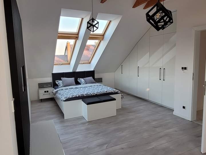 Pokój na poddaszu ze skośnym sufitem, łóżkiem z ciemnym zagłówkiem, szafkami nocnymi, białymi szafami na całej ścianie i dwoma oknami dachowymi z widokiem na dachy domów.