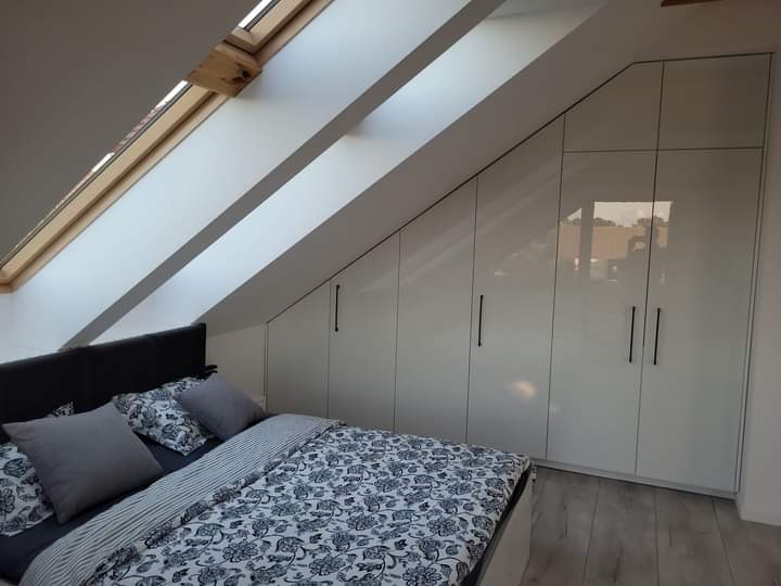 Sypialnia na poddaszu z zabudową szaf pod skosem, białe, lakierowane fronty, czarne uchwyty, łóżko z pościelą w kwiaty, okno dachowe.