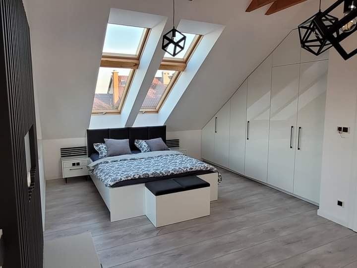 Pokój na poddaszu z łóżkiem, szafą w zabudowie pod skosem, oknami dachowymi i modernistycznymi lampami wiszącymi.