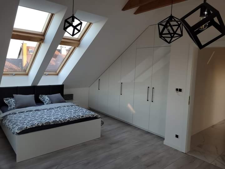 Jasna sypialnia na poddaszu ze skośnym sufitem, oknami dachowymi, białymi szafami w zabudowie i geometrycznymi lampami wiszącymi.