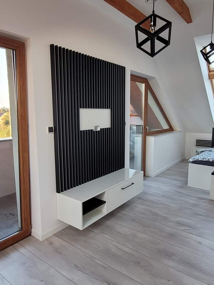 Nowoczesna aranżacja ściany z czarnymi lamelami i białą szafką RTV w pokoju na poddaszu ze skośnym oknem i drewnianymi belkami stropowymi, oświetlonym geometrycznymi lampami.