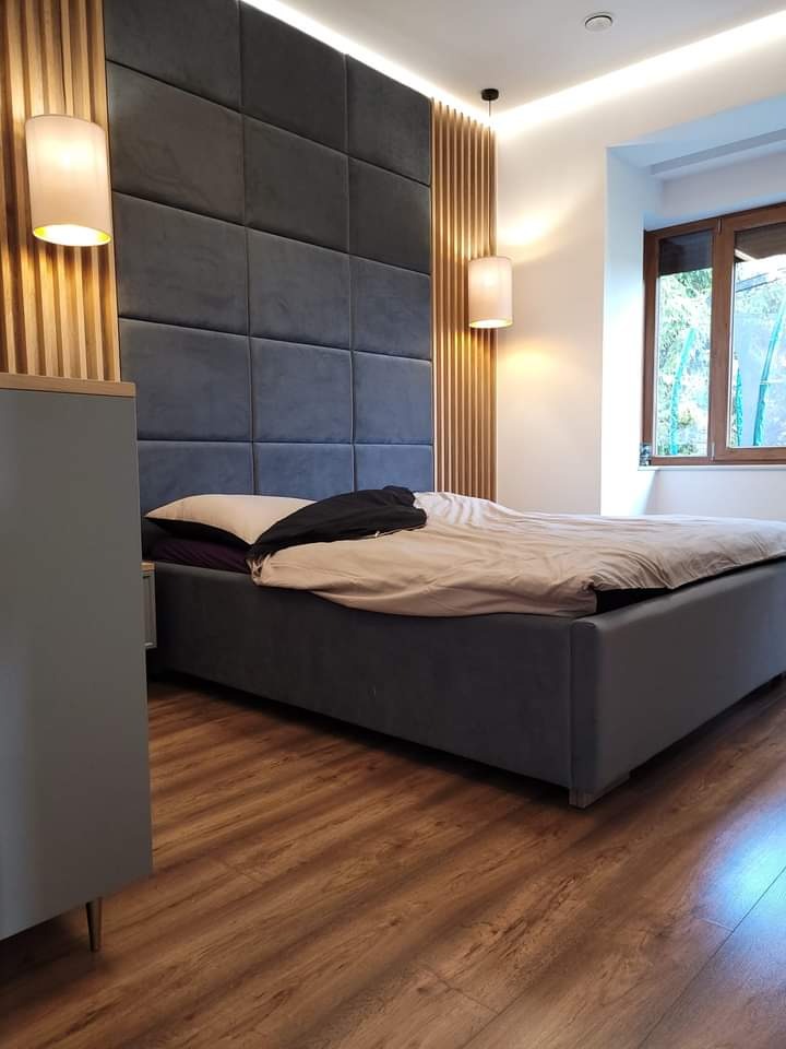 Sypialnia z łóżkiem kontynentalnym, tapicerowanym panelem ściennym w odcieniach szarości i drewnianymi lamelami, oświetlona lampami wiszącymi i kinkietami, z widokiem na zieleń za oknem.