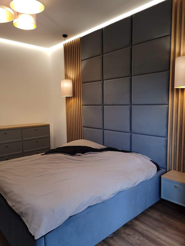 Nowoczesna sypialnia z łóżkiem kontynentalnym, wezgłowie z paneli tapicerowanych w kolorze grafitowym, oświetlenie LED podsufitowe i pionowe panele drewniane po bokach, szafka nocna i komoda...