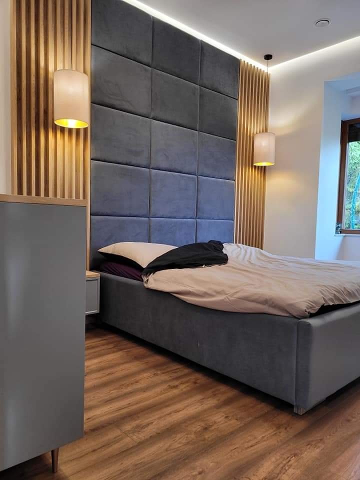 Sypialnia z łóżkiem z szarym tapicerowanym wezgłowiem z paneli, drewnianymi lamelami na ścianie i wiszącymi lampami z beżowymi abażurami. Widoczne okno z zielenią na zewnątrz.