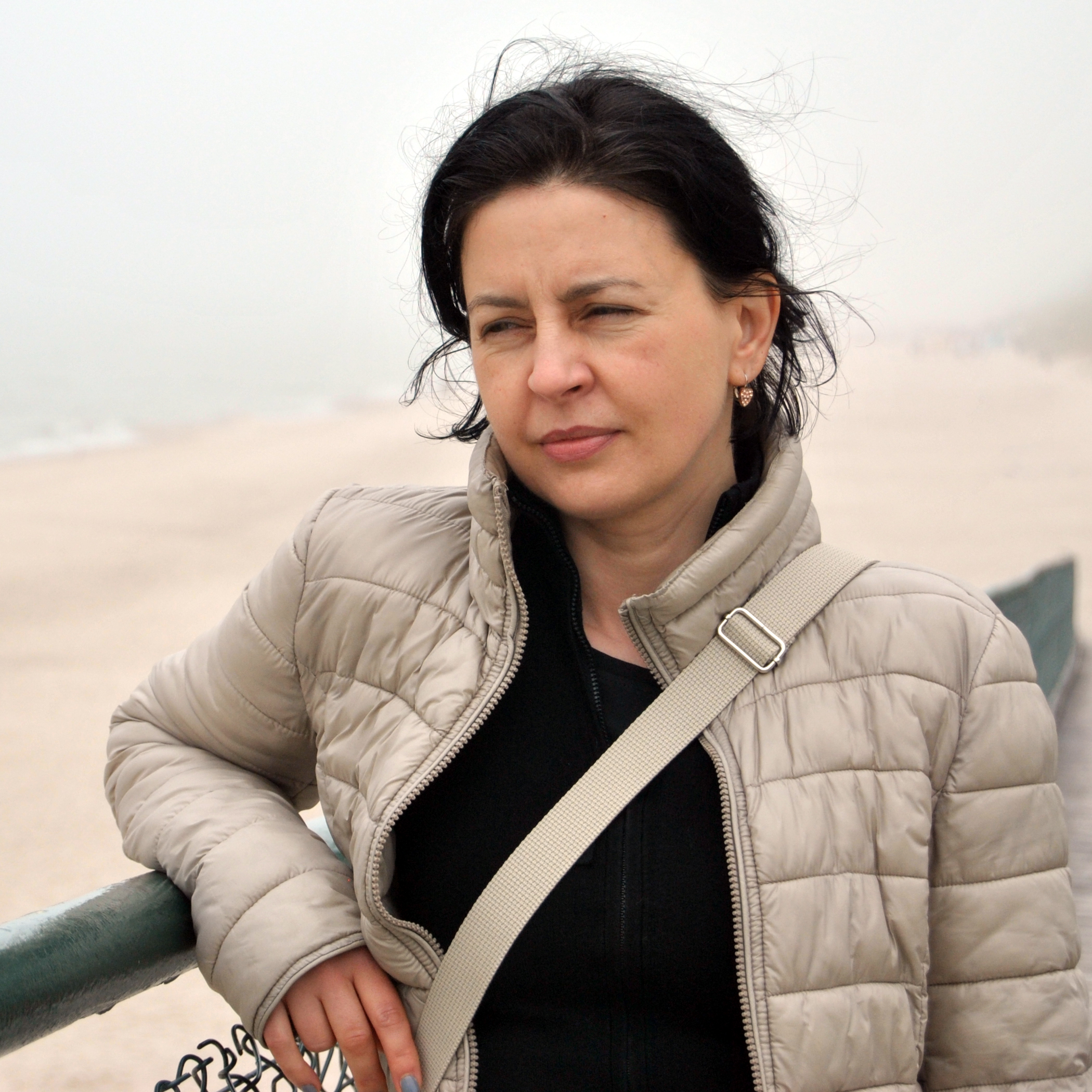 Zamyślona kobieta w beżowej kurtce, oparta o zieloną barierkę na tle mglistej plaży, z torbą przewieszoną przez ramię.