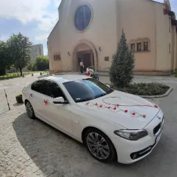 Białe BMW udekorowane czerwonymi wstążkami i kwiatami w kształcie serc, zaparkowane przed kościołem z osobami w tle.