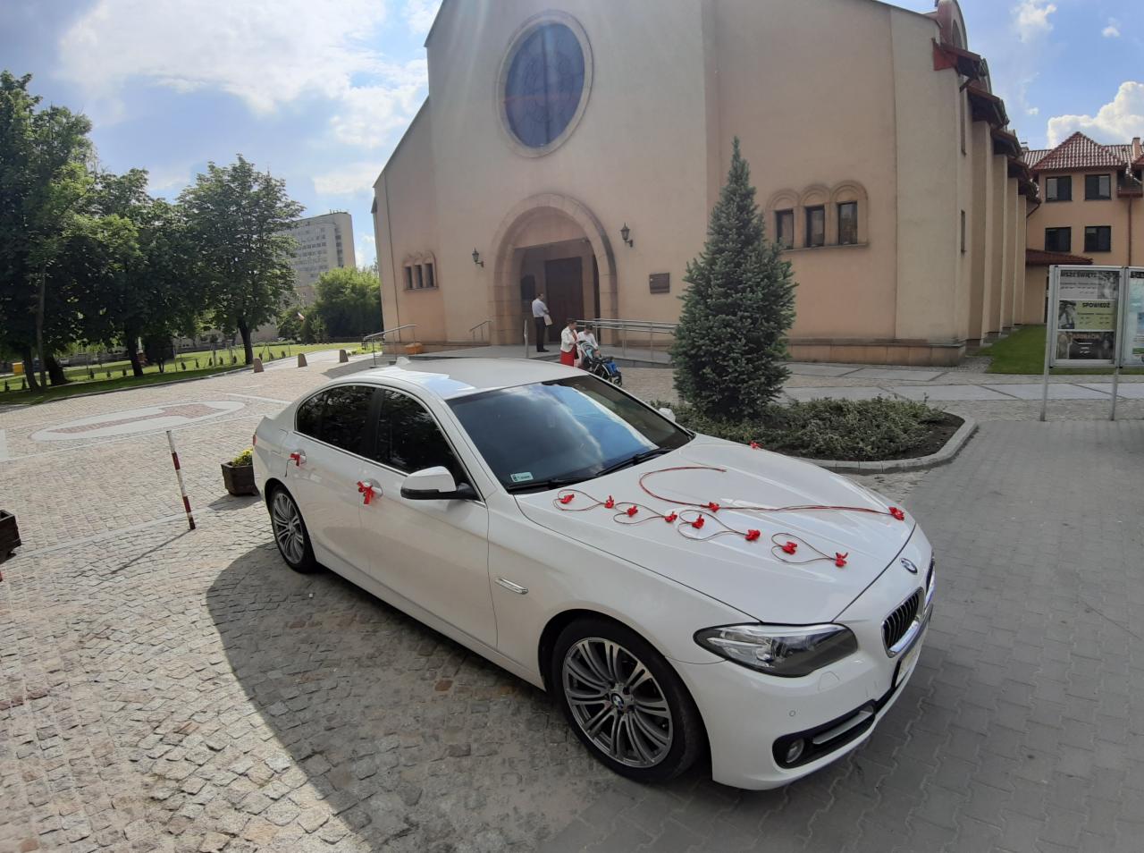 Białe BMW udekorowane czerwonymi wstążkami i kwiatami w kształcie serc, zaparkowane przed kościołem z osobami w tle.