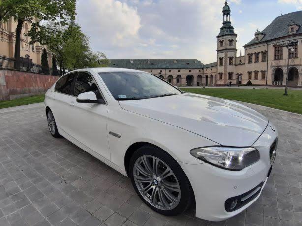 Biała limuzyna BMW serii 5 zaparkowana na brukowanej nawierzchni z zabytkowym budynkiem w tle, widoczne detale felg i reflektorów.