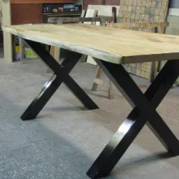 Stół z blatem z jasnego drewna i czarnymi, metalowymi nogami w kształcie litery X, widoczne w warsztacie.