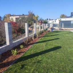 Ogrody TG Tarnowskie Góry - Układanie kory i sadzenie roślin wzdłuż ogrodzenia z betonowych bloczków na trawniku. Pracownik w trakcie pracy na tle zabudowy jednorodzinnej.