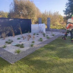Ogrody TG Tarnowskie Góry - Nowoczesny ogród z białym żwirem i ozdobnymi trawami, zabezpieczone krzewy, ogrodnik z dmuchawą do liści, zielony trawnik i ciemne ogrodzenie w tle.