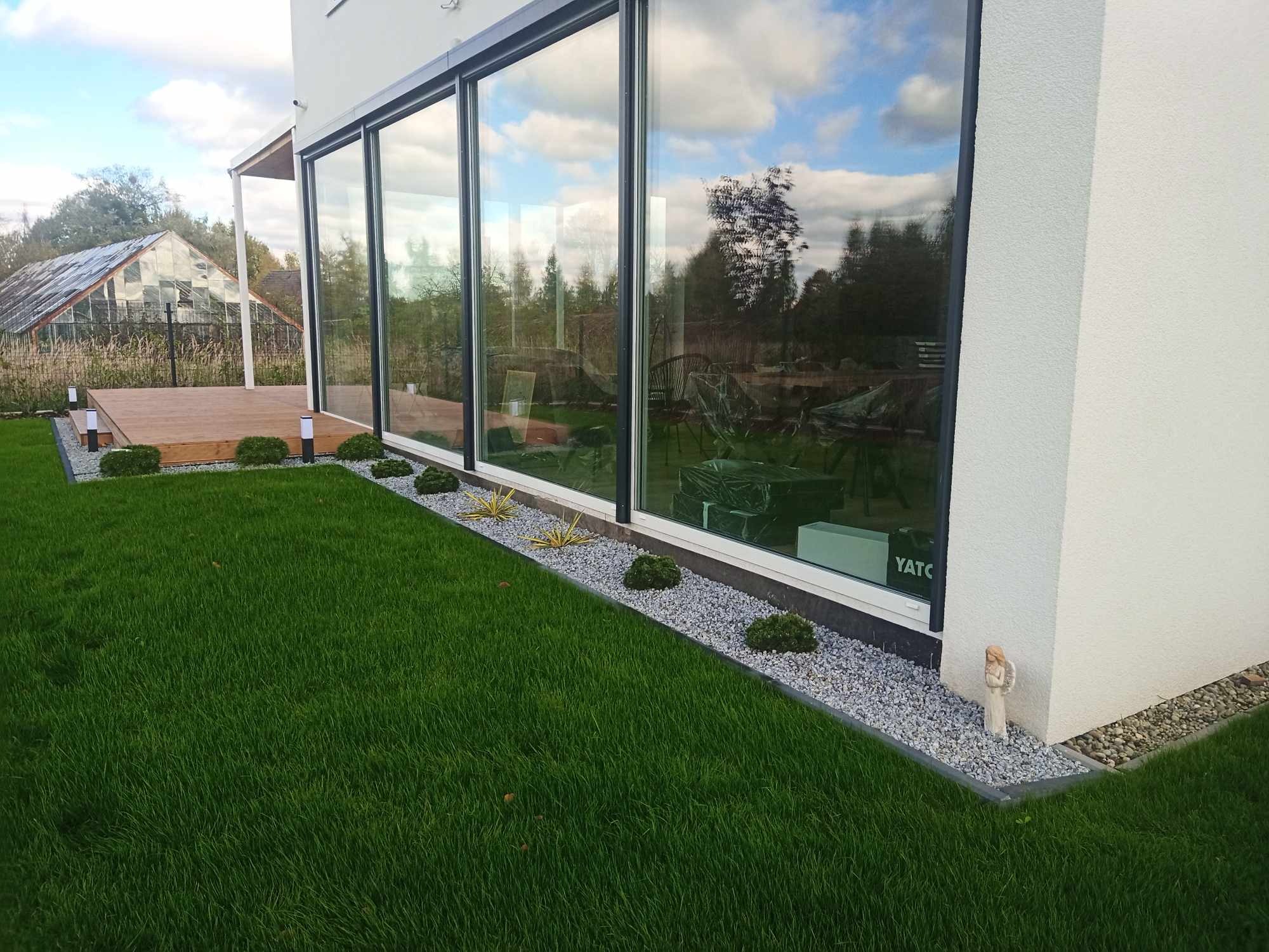 Nowoczesny ogród przydomowy z równo przystrzyżonym trawnikiem, ozdobnymi krzewami i szarą rabatą żwirową wzdłuż ściany budynku z dużymi oknami, w których odbija się błękitne niebo i zieleń drzew.