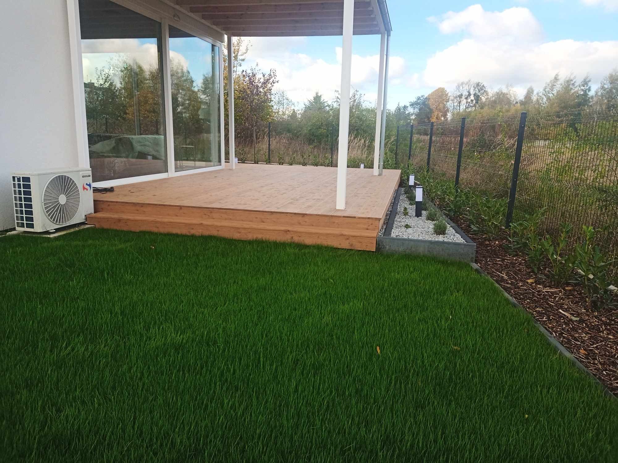 Nowoczesny taras z drewnianą podłogą i schodkami, otoczony intensywnie zieloną trawą, z widokiem na ogrodzenie i roślinność w tle.