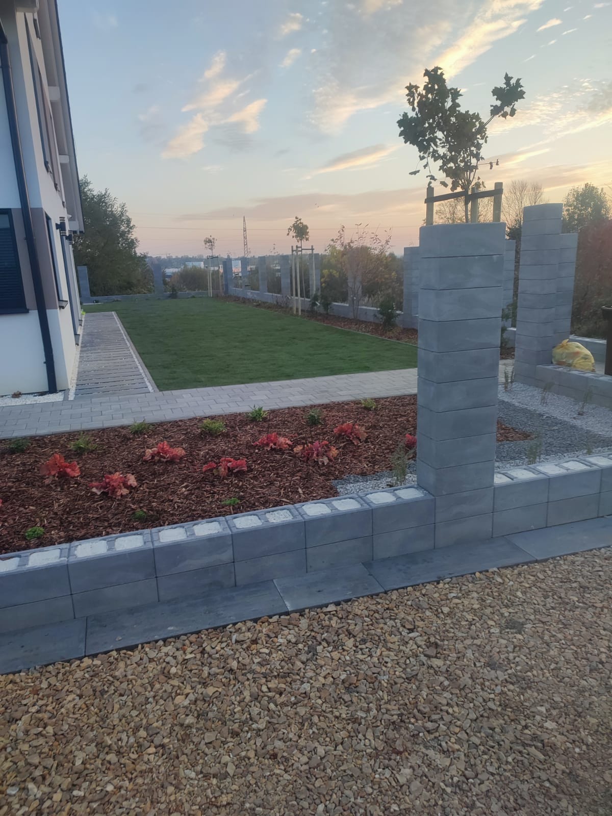 Nowoczesny ogród z równo przystrzyżonym trawnikiem, rabatami z korą i czerwonymi roślinami, oraz szarą obudową z betonowych bloczków. Widok na dom i drzewa.