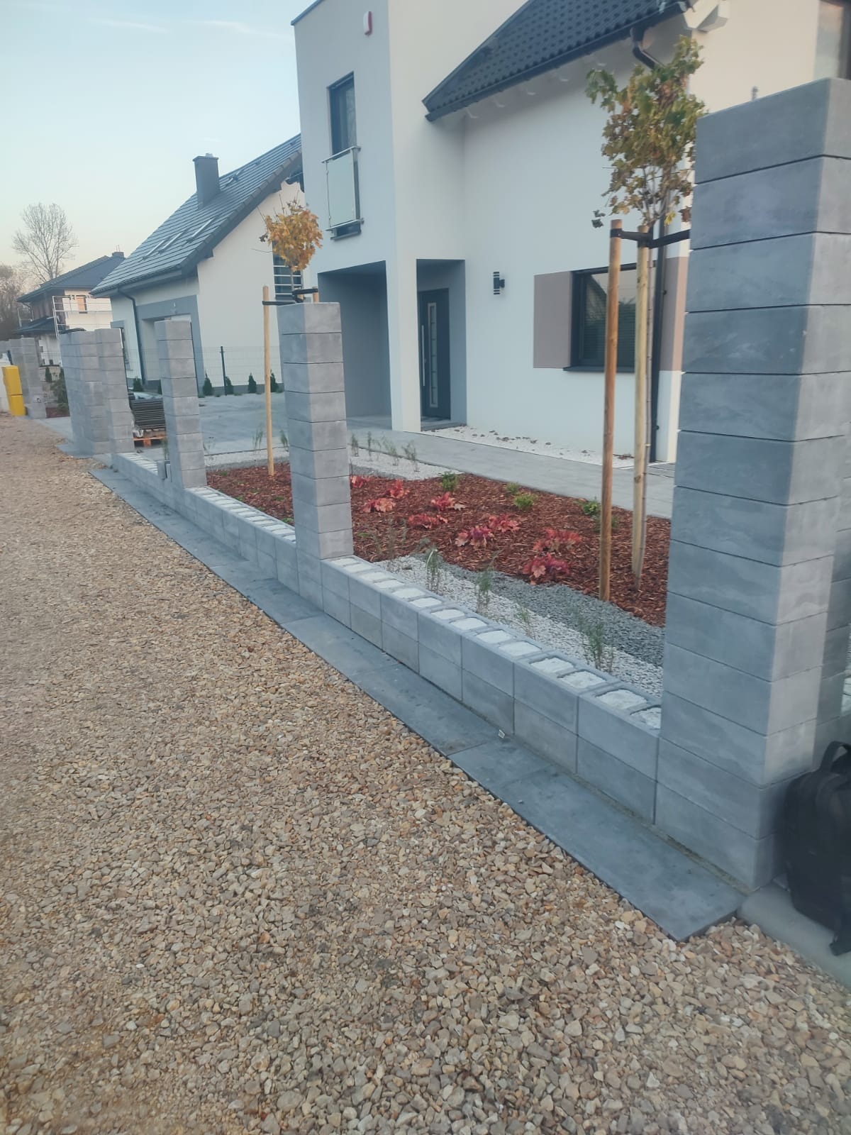 Nowoczesny ogródek przed domem z szarej kostki, wypełniony korą i roślinami. Widoczne słupki i obrzeża z kostki, żwirowe podłoże. Minimalistyczny design.