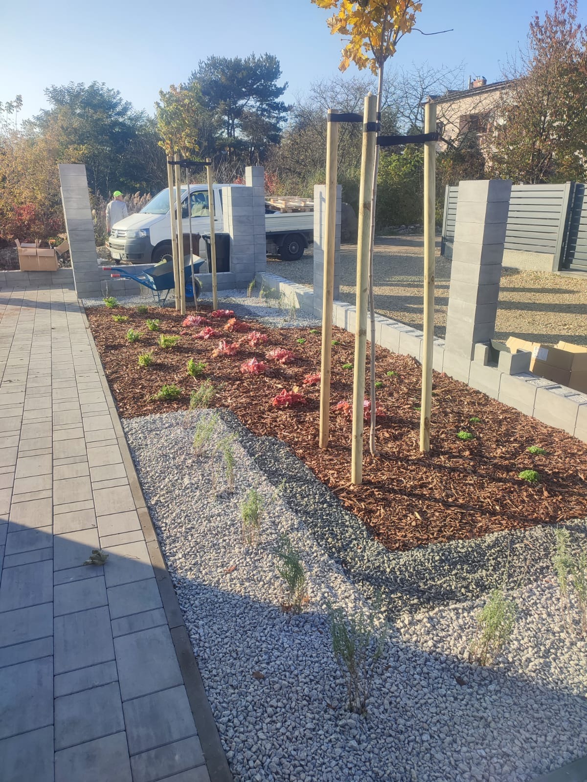 Świeżo założony ogród z drzewkami podpartymi drewnianymi palikami, rabatami wysypanymi korą i kamieniami, obrzeża z szarych bloczków.