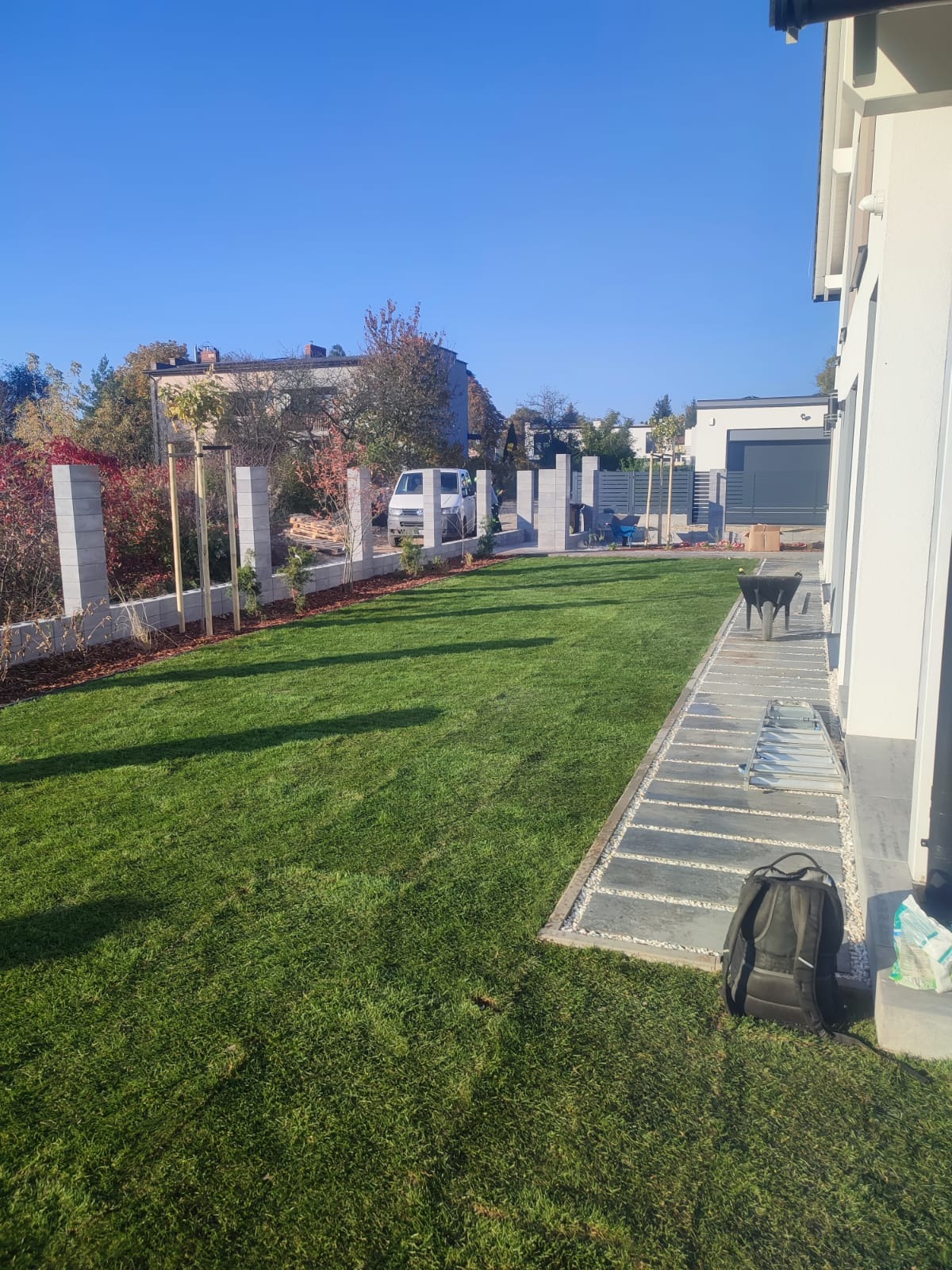 Ogród po założeniu: świeża trawa, obrzeża z kory, ścieżka z płyt kamiennych wzdłuż budynku, nowe nasadzenia drzew i krzewów, nowoczesne ogrodzenie z elementami betonowymi.