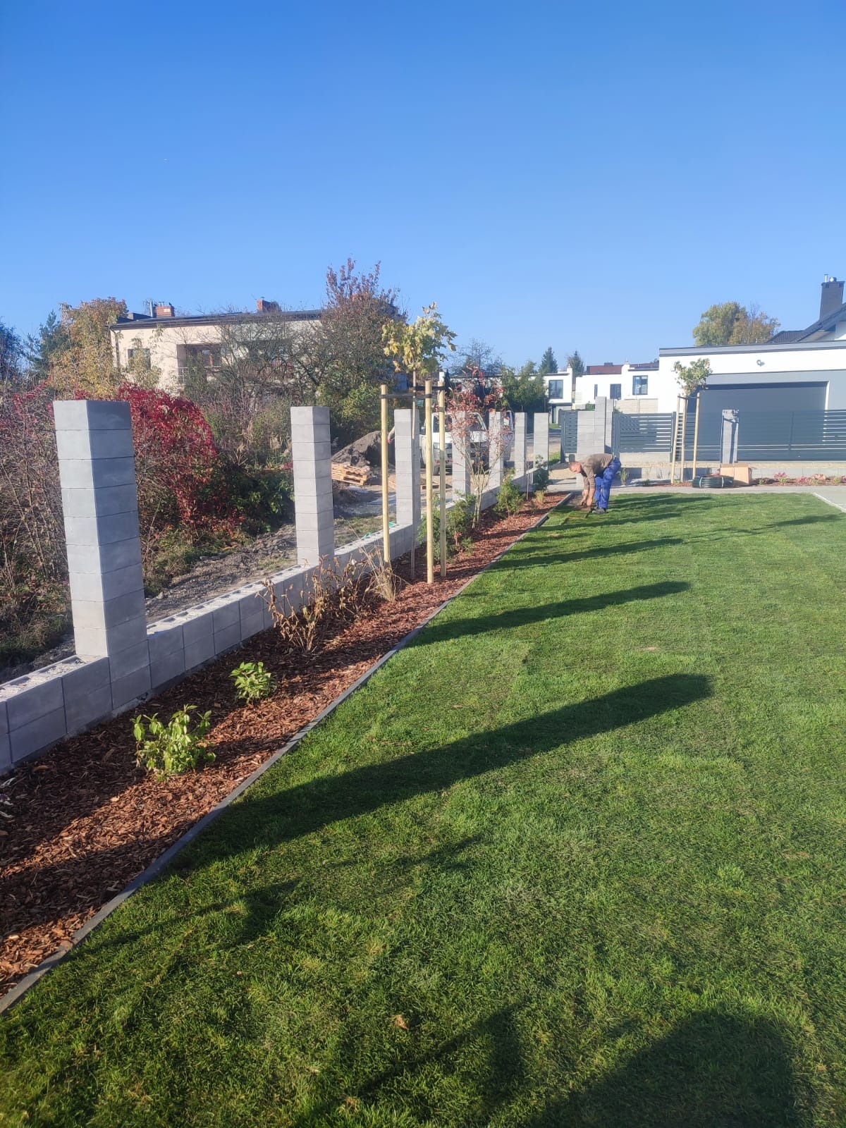 Układanie kory i sadzenie roślin wzdłuż ogrodzenia z betonowych bloczków na trawniku. Pracownik w trakcie pracy na tle zabudowy jednorodzinnej.