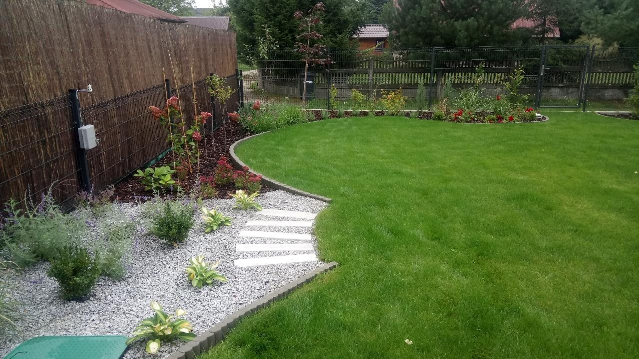 Uporządkowany ogród z trawnikiem, ścieżką z jasnych płyt i rabatami z krzewami oraz kwiatami, otoczony płotem z siatki i bambusową osłoną.