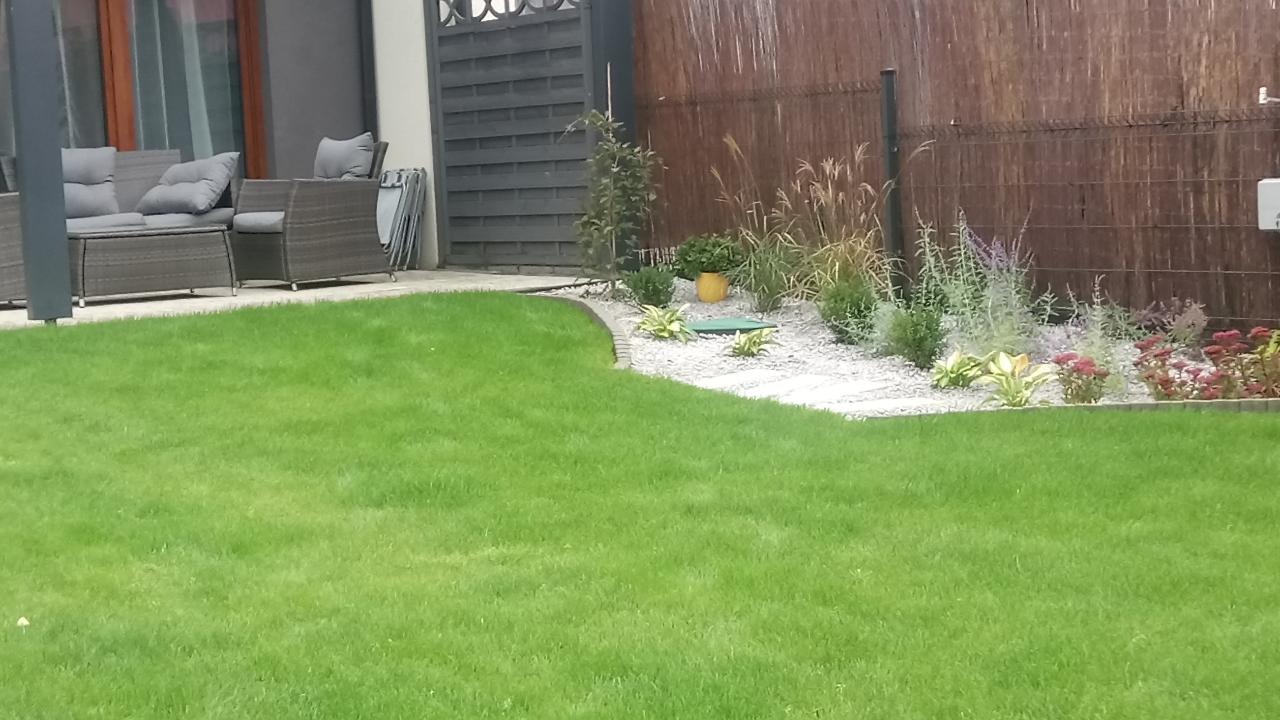 Zadbany trawnik z obrzeżem i rabatą z białym żwirem, ozdobnymi trawami i roślinami. W tle meble ogrodowe i płot z brązowych paneli.