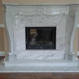 Elegancki, marmurowy portal kominkowy z czarnym wkładem, wzór z subtelnymi zdobieniami, wykończenie w odcieniach bieli i szarości.