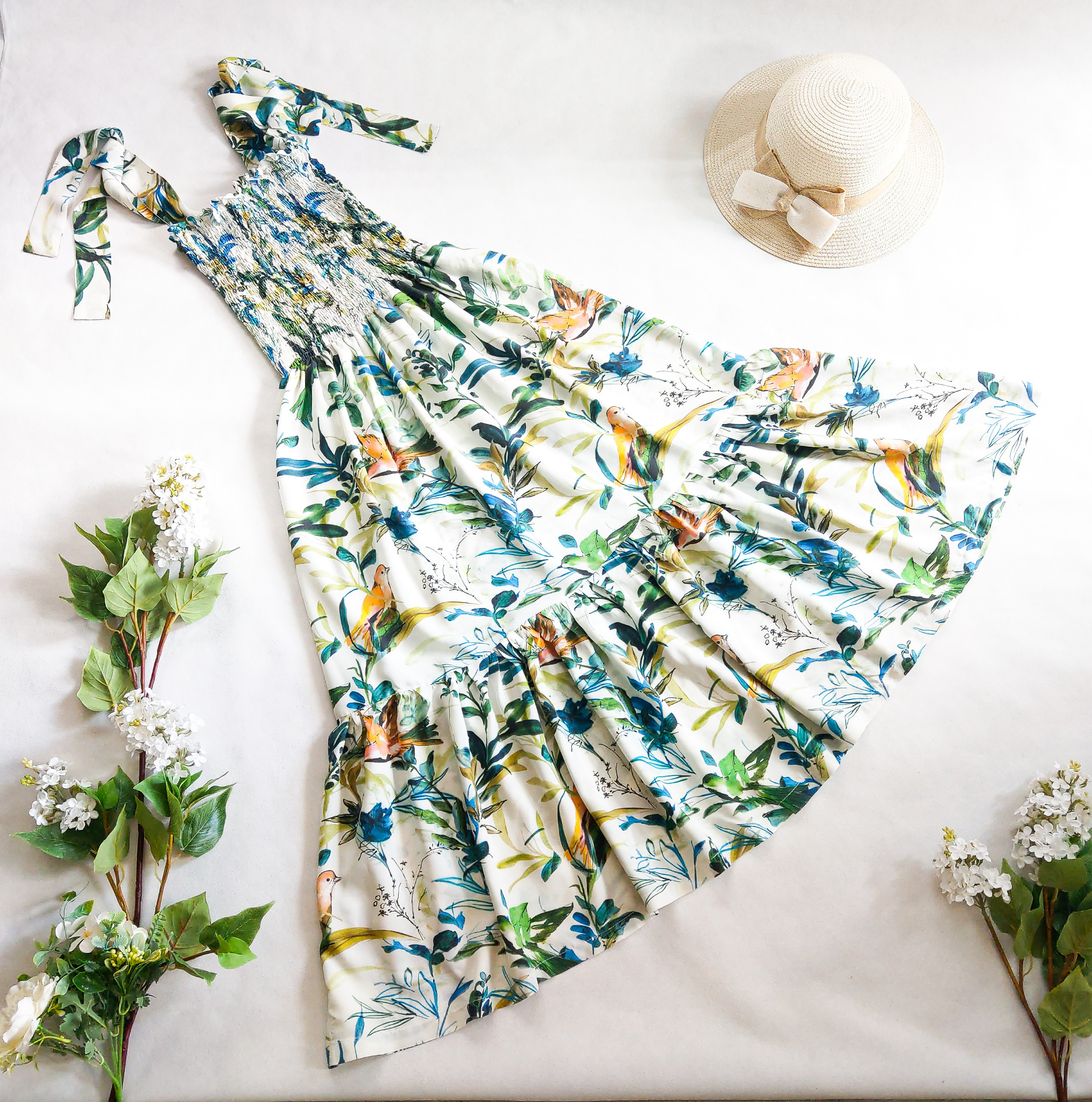 Letnia sukienka z motywem ptaków i roślin, leżąca obok słomkowego kapelusza i gałązek kwitnącego bzu na białym tle.