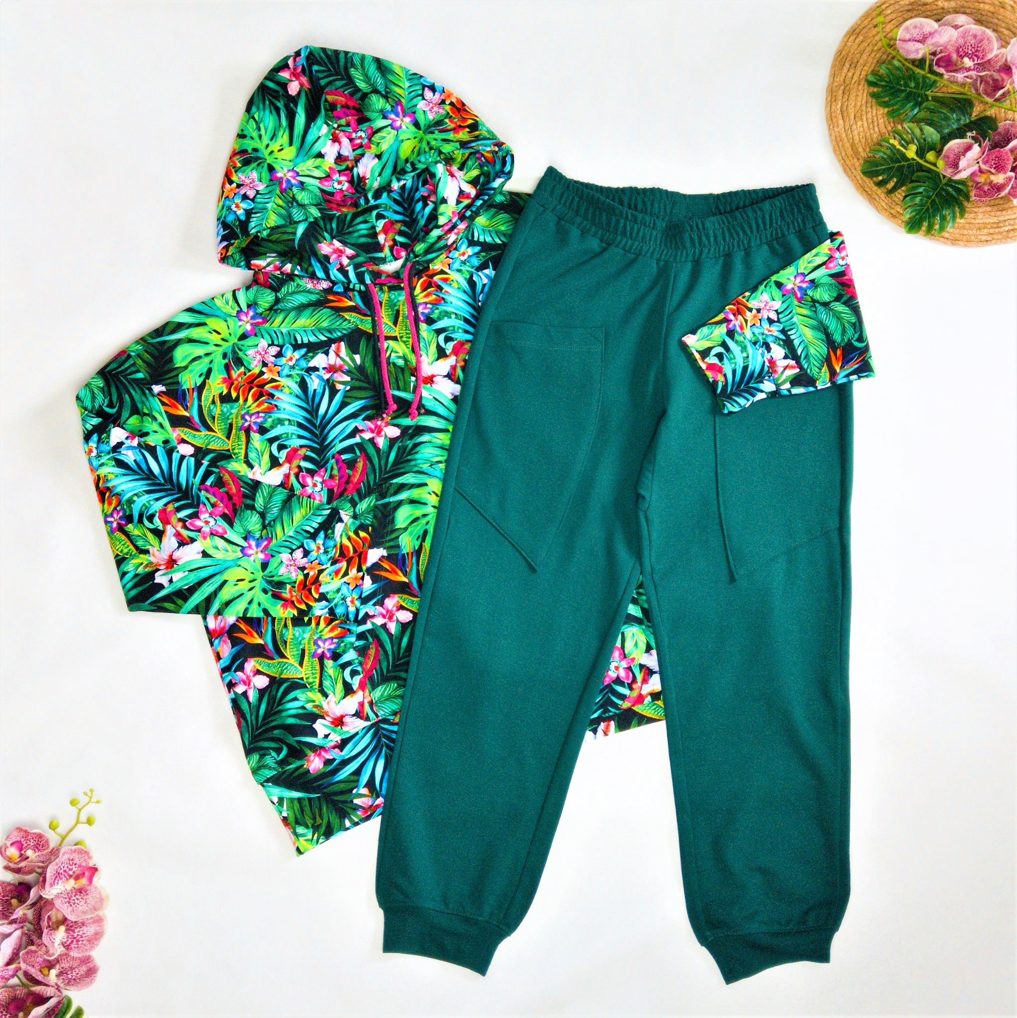 Komplet dresowy: bluza z kapturem w tropikalny wzór i gładkie, zielone spodnie dresowe, ułożone na białym tle z dekoracją kwiatową.