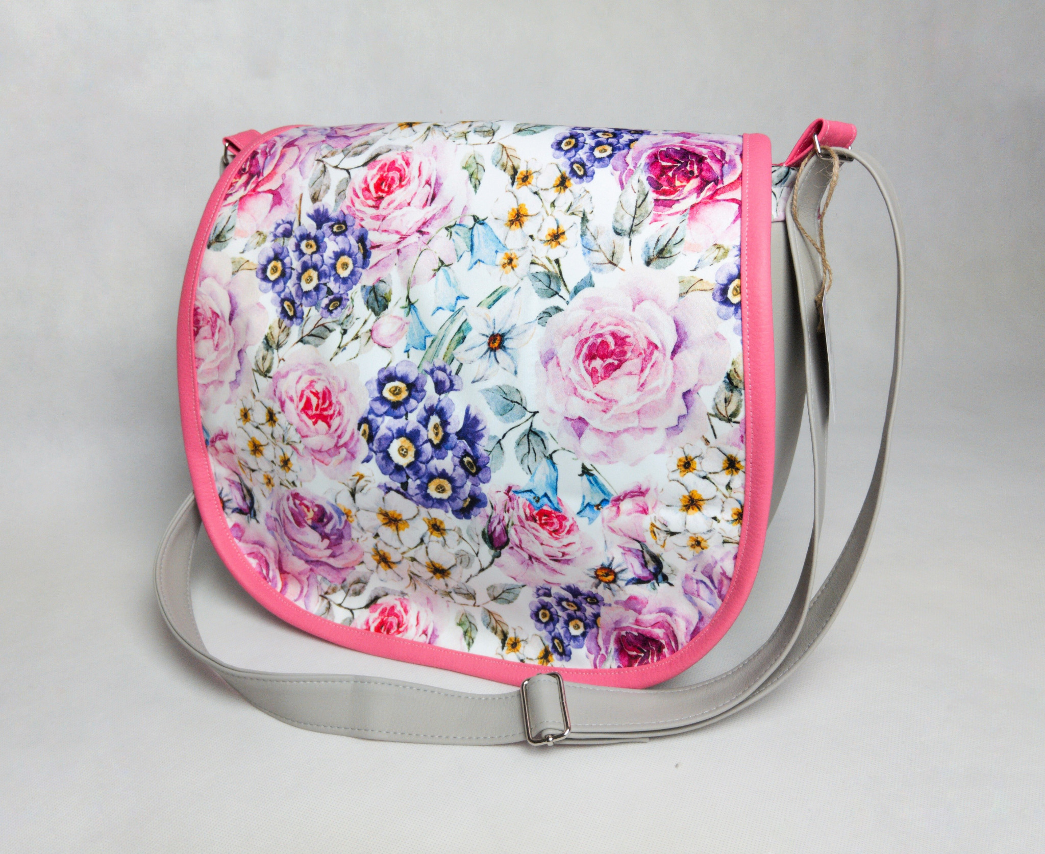 Torba listonoszka z kwiatowym wzorem w odcieniach różu, fioletu i błękitu, obszyta różową lamówką i z szarym paskiem, leżąca na jasnym tle.