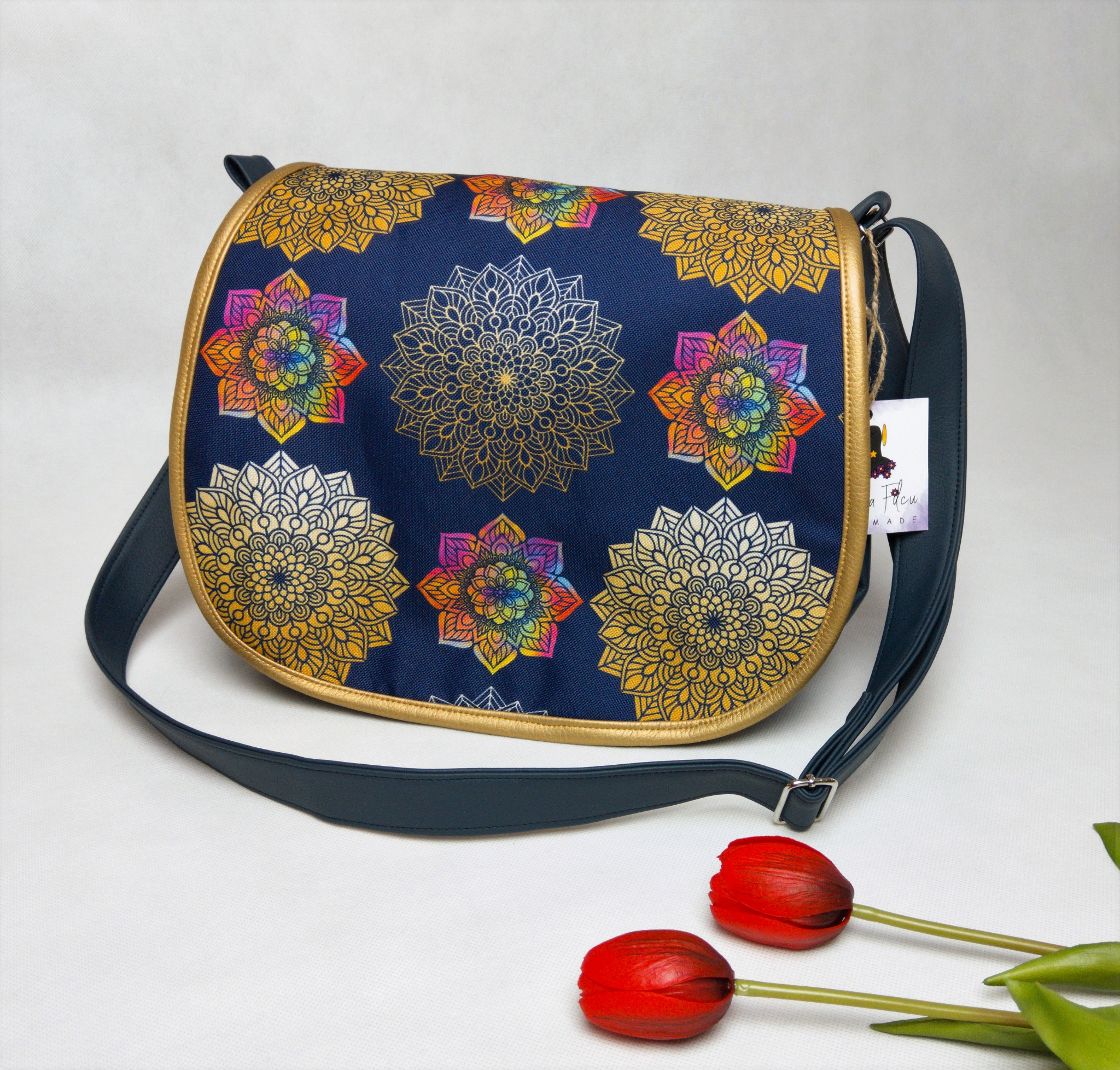 Damska torebka listonoszka z granatowego materiału w złote mandale i tęczowe kwiaty, wykończona złotą lamówką, leżąca na białym tle obok dwóch czerwonych tulipanów.