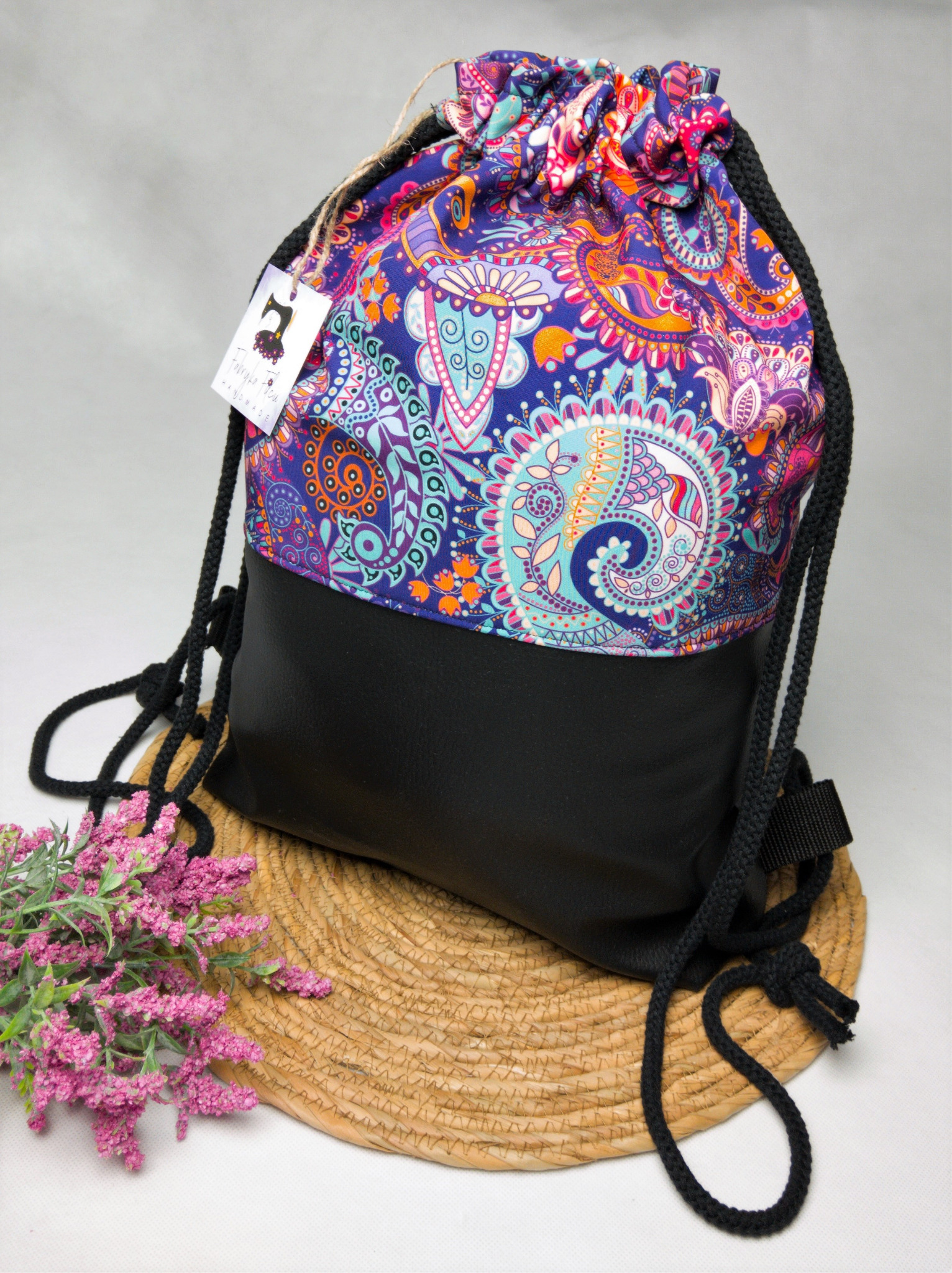 Plecak-worek z czarnego materiału i kolorowej tkaniny w orientalne wzory, na słomianej podkładce z dodatkiem dekoracyjnych, różowych kwiatów i metką.
