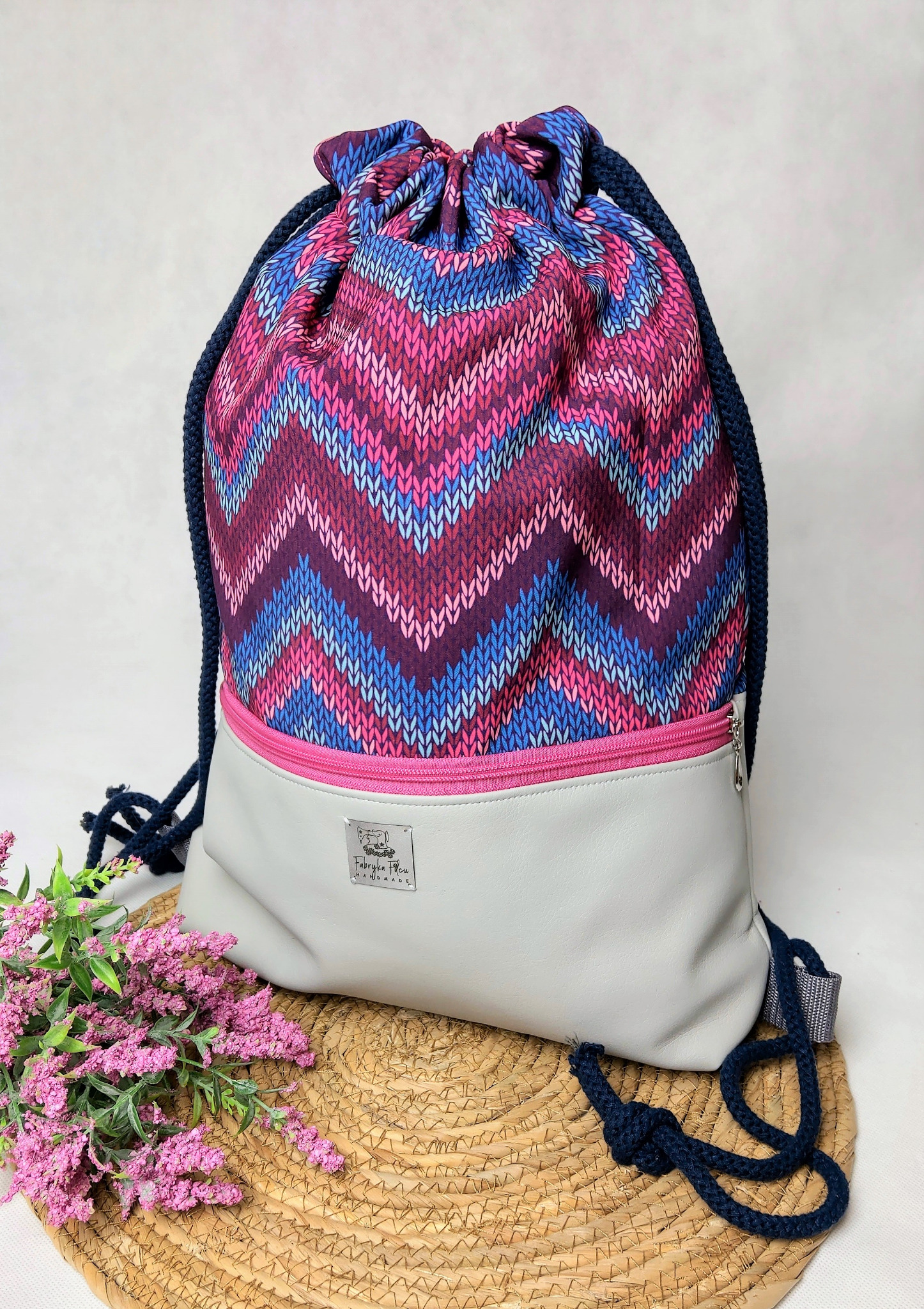 Wielobarwny plecak-worek ze ściągaczem i kieszenią na zamek, z etykietą 'Fabryka Filcu Handmade', ustawiony na słomianej podkładce z dekoracją z różowych kwiatów.