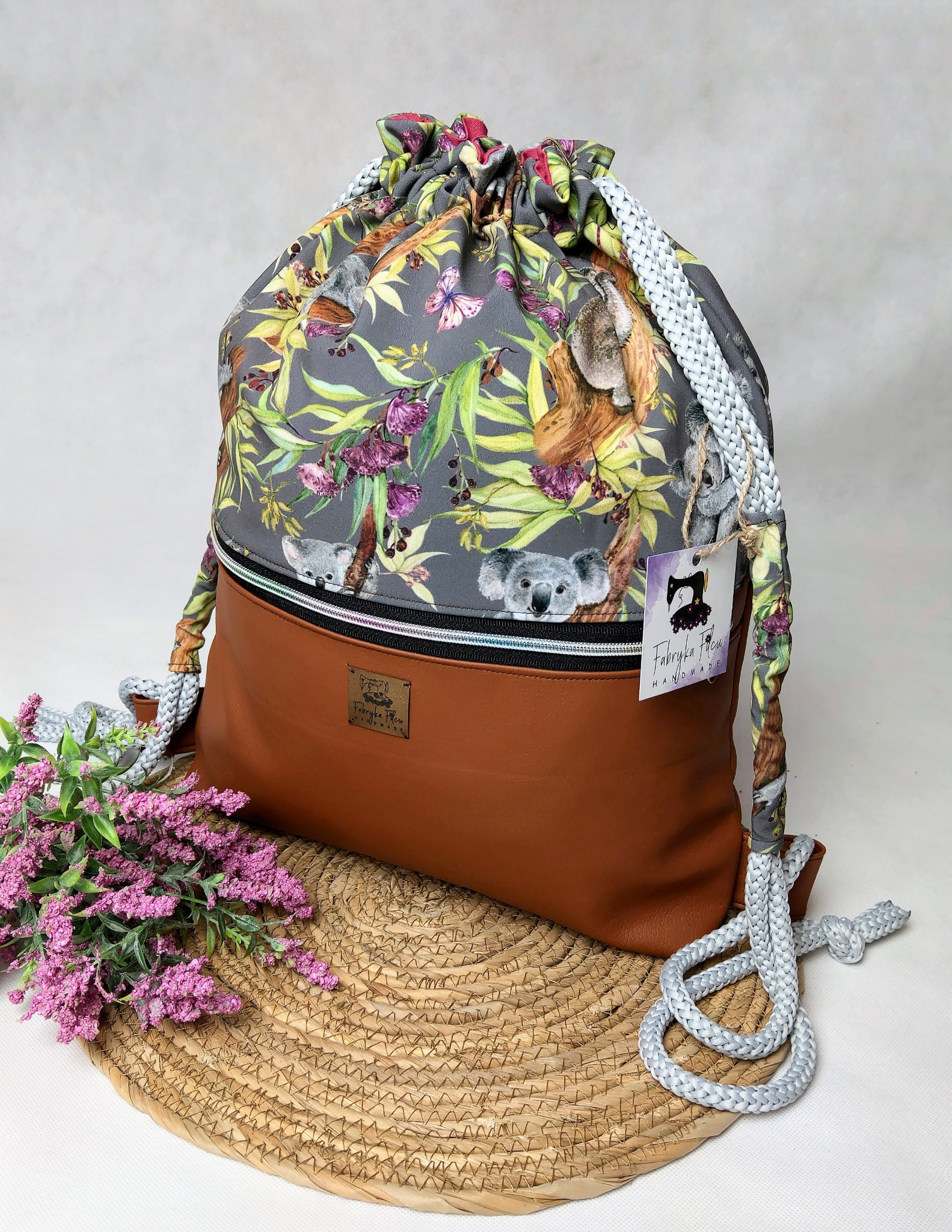 Plecak-worek z brązowej skóry ekologicznej i szarej tkaniny z motywem koali, kwiatów i motyli, stojący na słomianej podkładce obok gałązki wrzosu, z widoczną metką producenta.