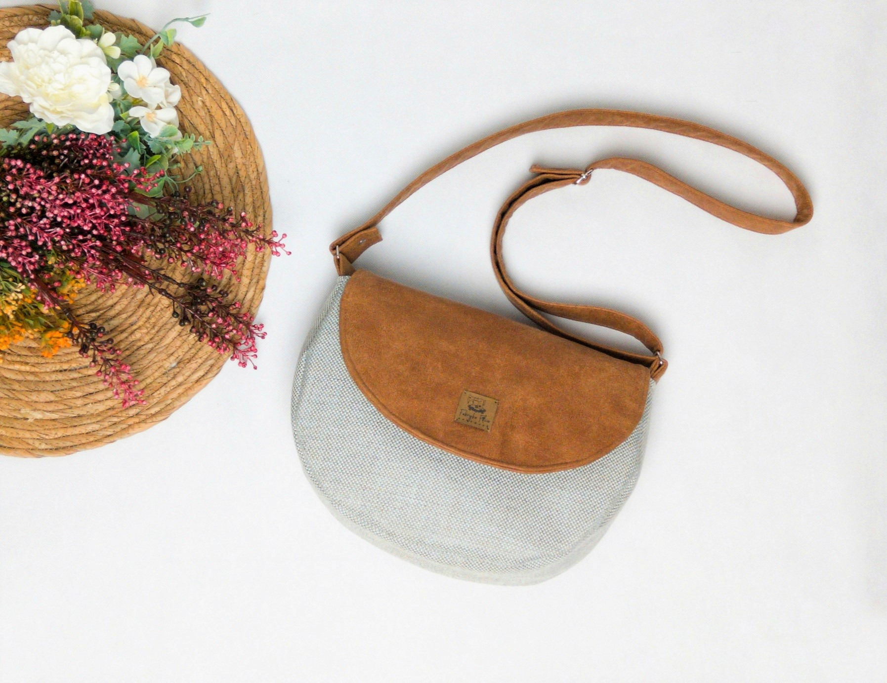 Beżowa torebka damska z zamszową klapą, leżąca obok dekoracyjnego wieńca z kwiatami na białym tle.