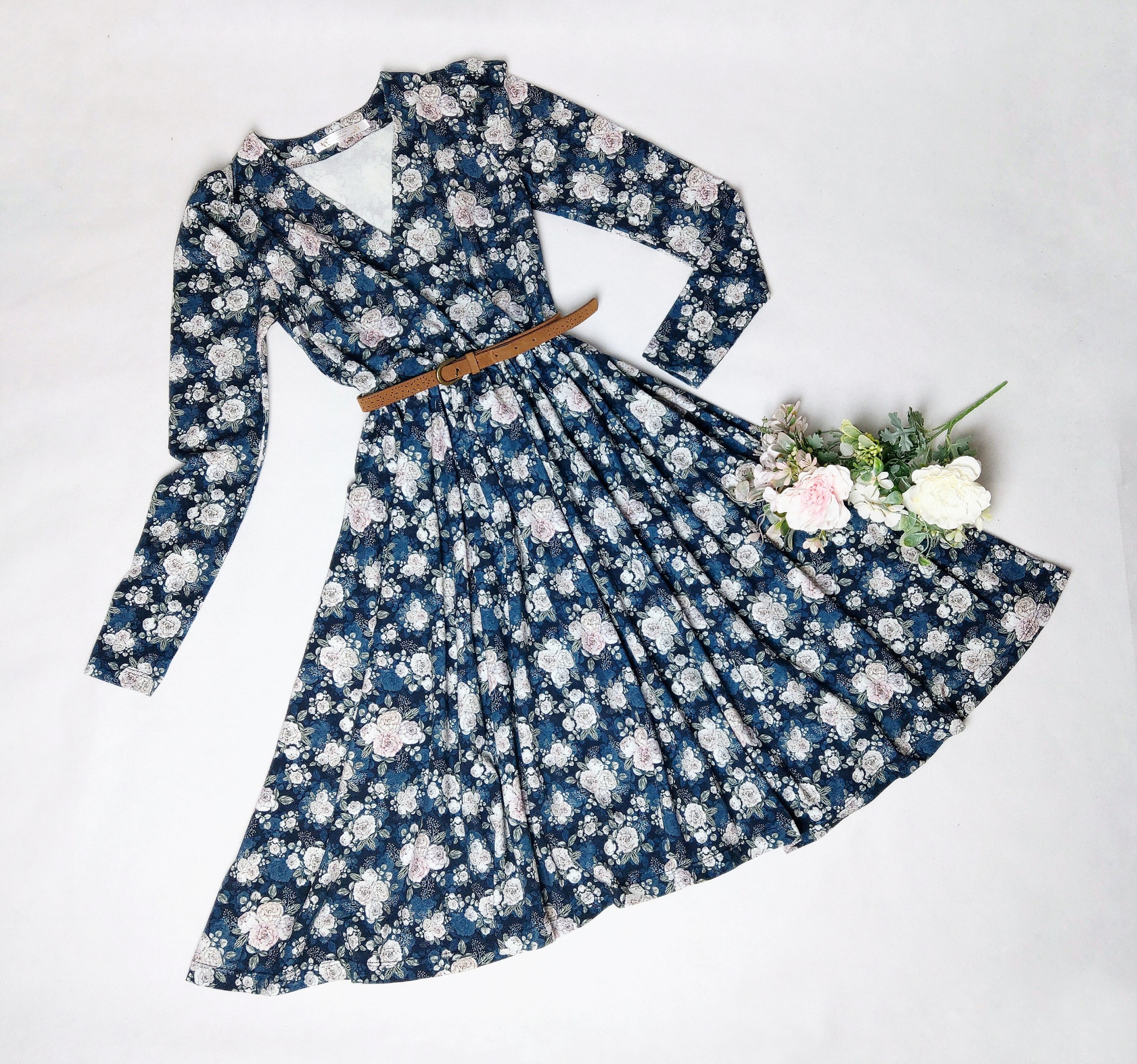 Damska sukienka z długim rękawem w kwiatowy wzór, przewiązana brązowym paskiem, leżąca na białym tle obok bukietu kwiatów.