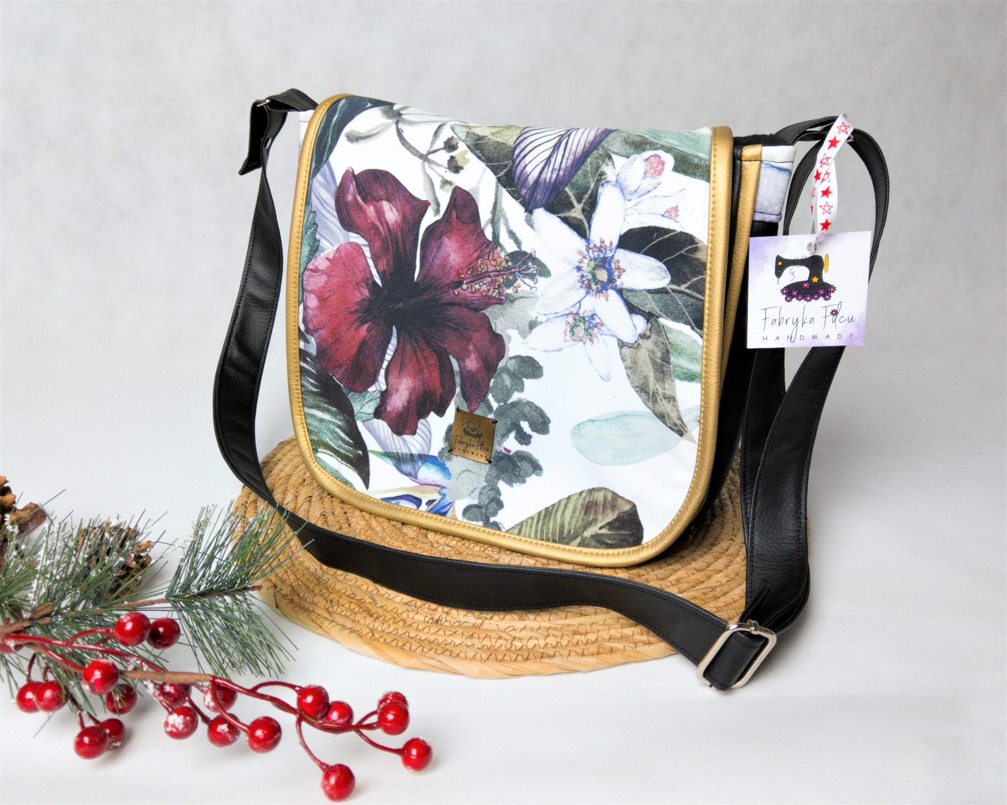 Torba damska typu listonoszka z motywem kwiatowym, czarnymi paskami i złotym obszyciem, prezentowana na plecionym podkładzie z dodatkiem świątecznej dekoracji.