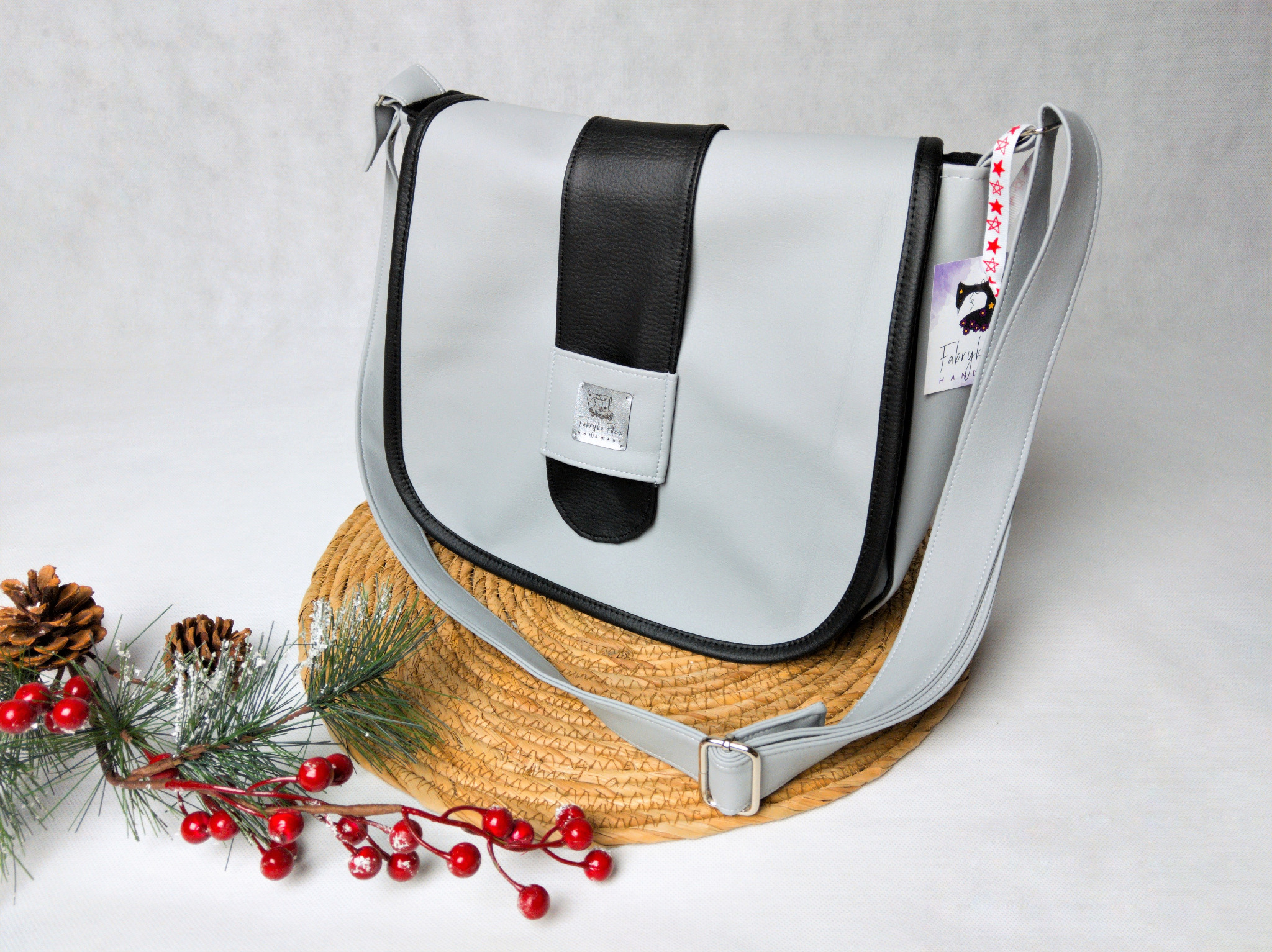 Elegancka, szaro-czarna torebka damska z paskiem, umieszczona na słomianej podkładce z dekoracją świąteczną z gałązek i czerwonych jagód, prezentowana na białym tle.