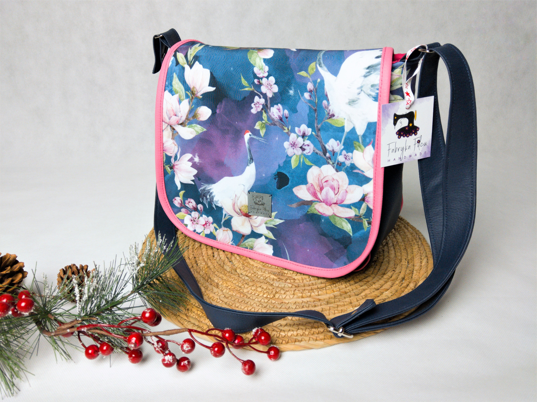 Torba listonoszka z motywem żurawi i magnolii na niebieskim tle, wykończona różową lamówką, leży na słomianej podkładce obok gałązki z czerwonymi jagodami i szyszek.