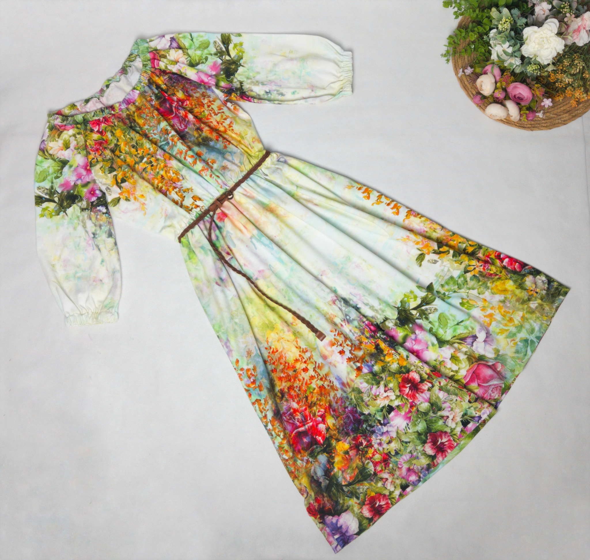 Letnia sukienka z długim rękawem, z motywem kwiatowym, leżąca na białym tle obok koszyka z kwiatami.