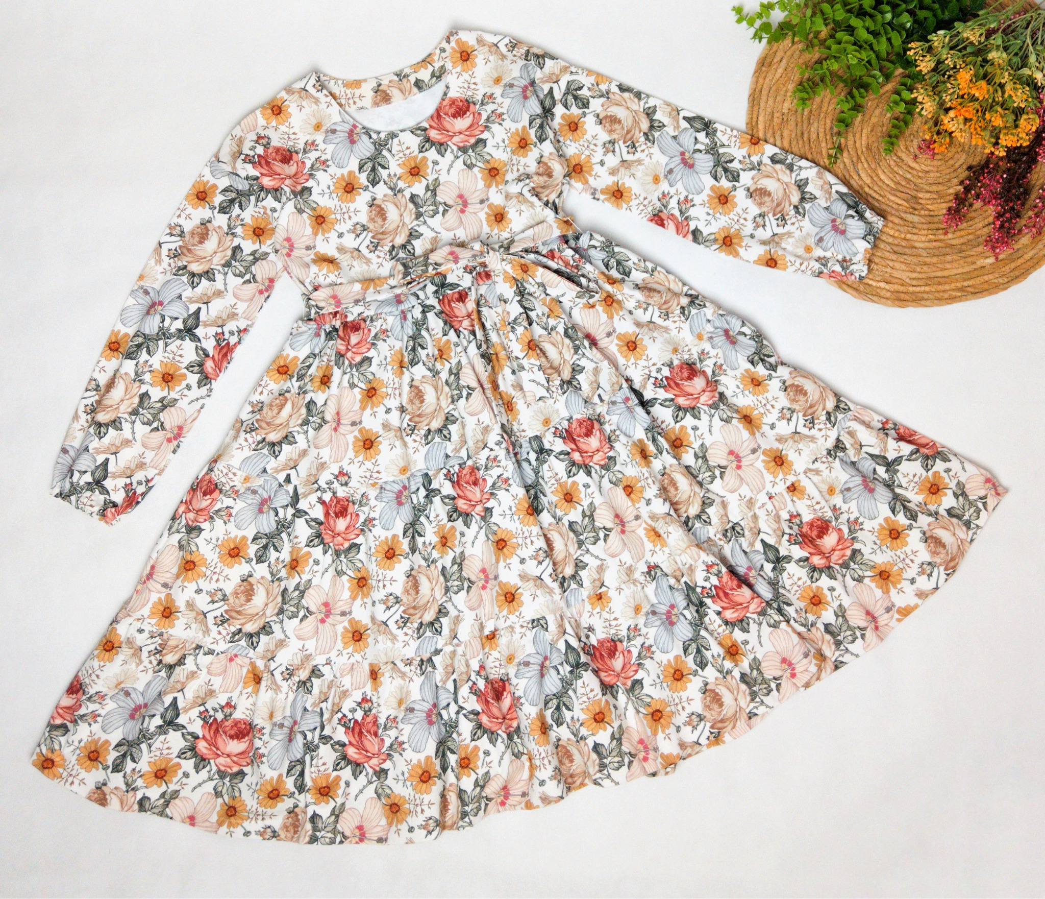 Dziewczęca sukienka z długim rękawem w kwiatowy wzór na białym tle, obok słomkowy kapelusz i dekoracja roślinna.
